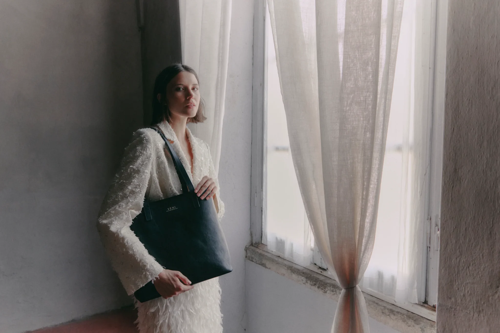 Maison Veni campaign, Milan: a model by a window holds the Rebekah tote in black leather