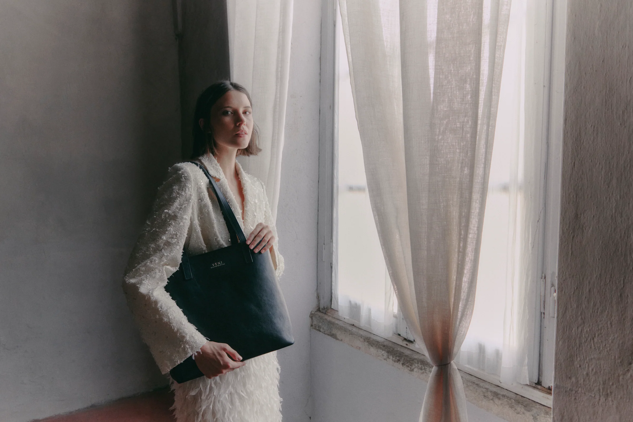 Maison Veni campaign, Milan: a model by a window holds the Rebekah tote in black leather
