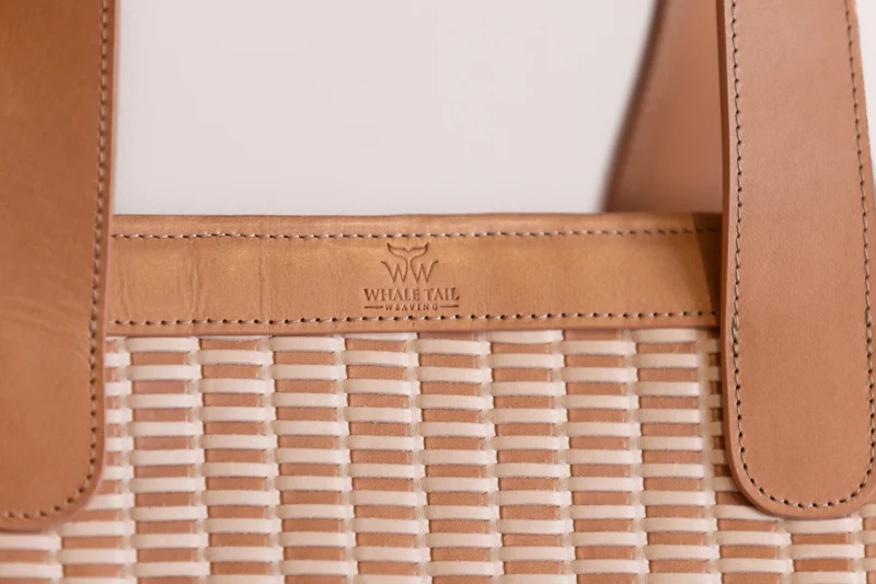 Close-up of the tote's leather handle base and embossed logo over the two-tone weave