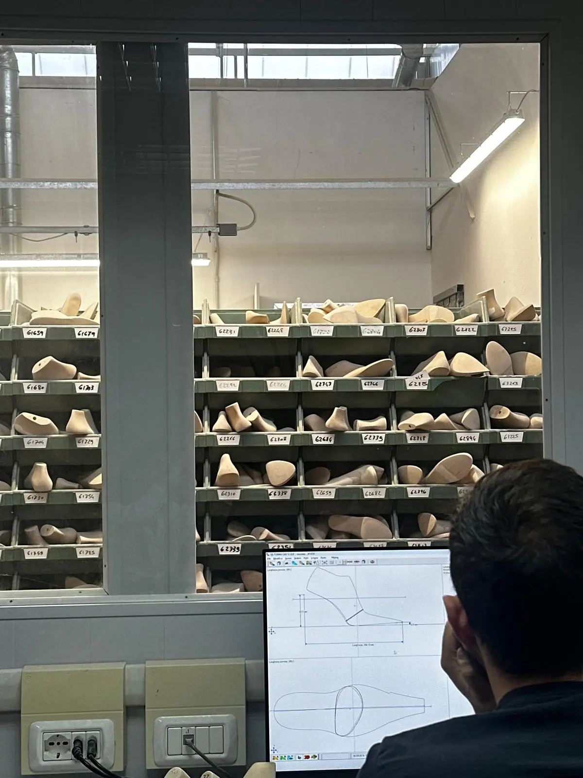 Shelves of wooden shoe lasts in the last maker's workshop, a technician at a drawing screen in the foreground