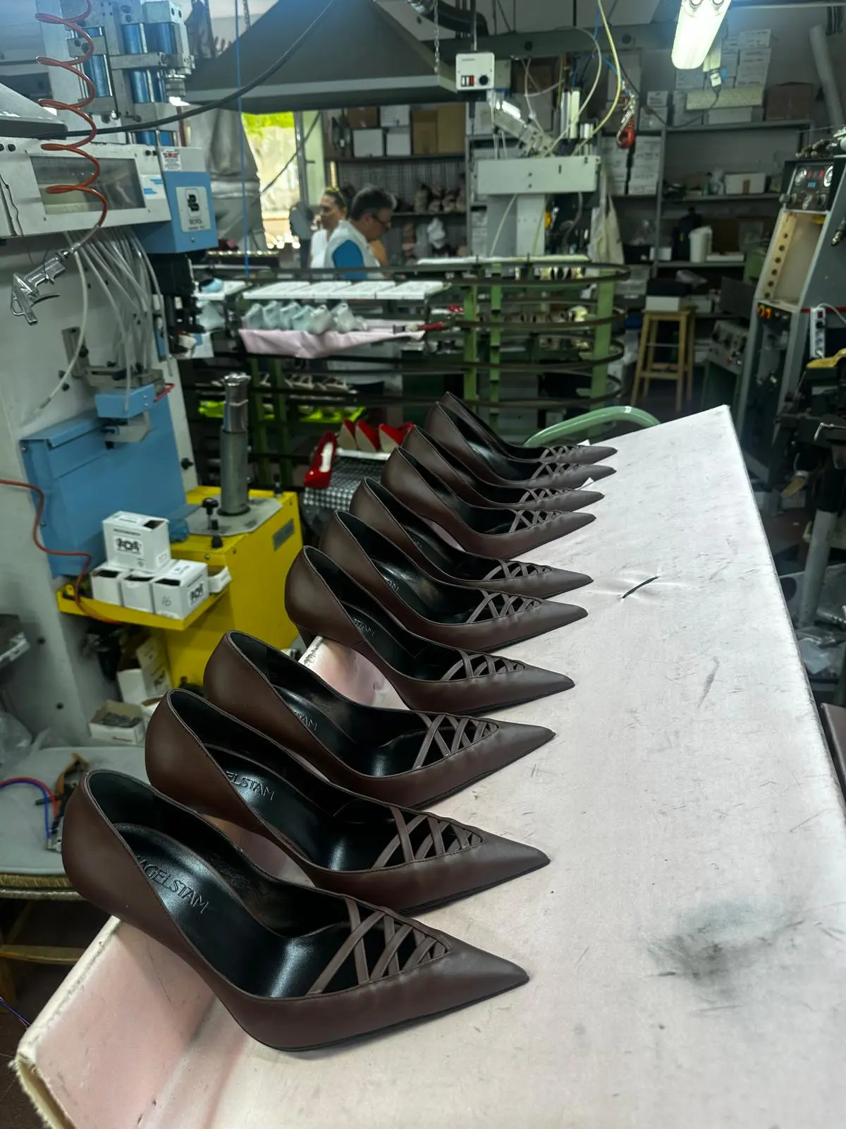 An artisan in a blue apron works on a line of brown pumps at a bench in the atelier