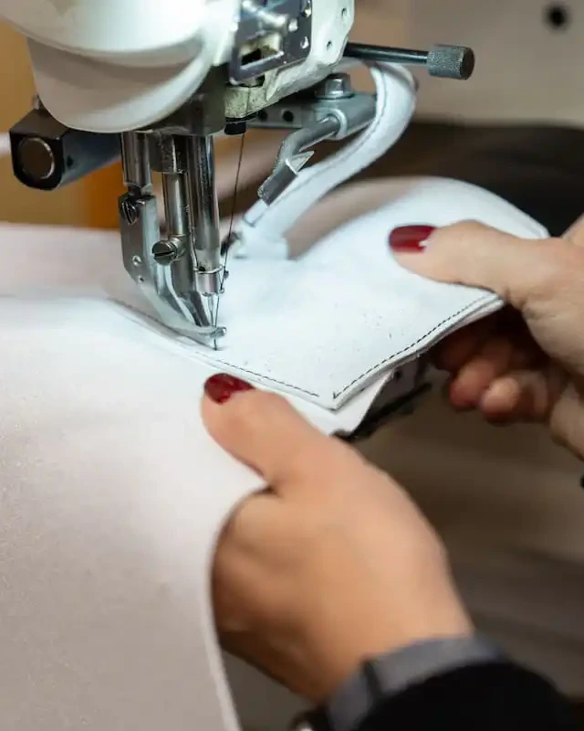 An artisan's hands guiding white leather under the needle of a sewing machine in an Italian handbag workshop